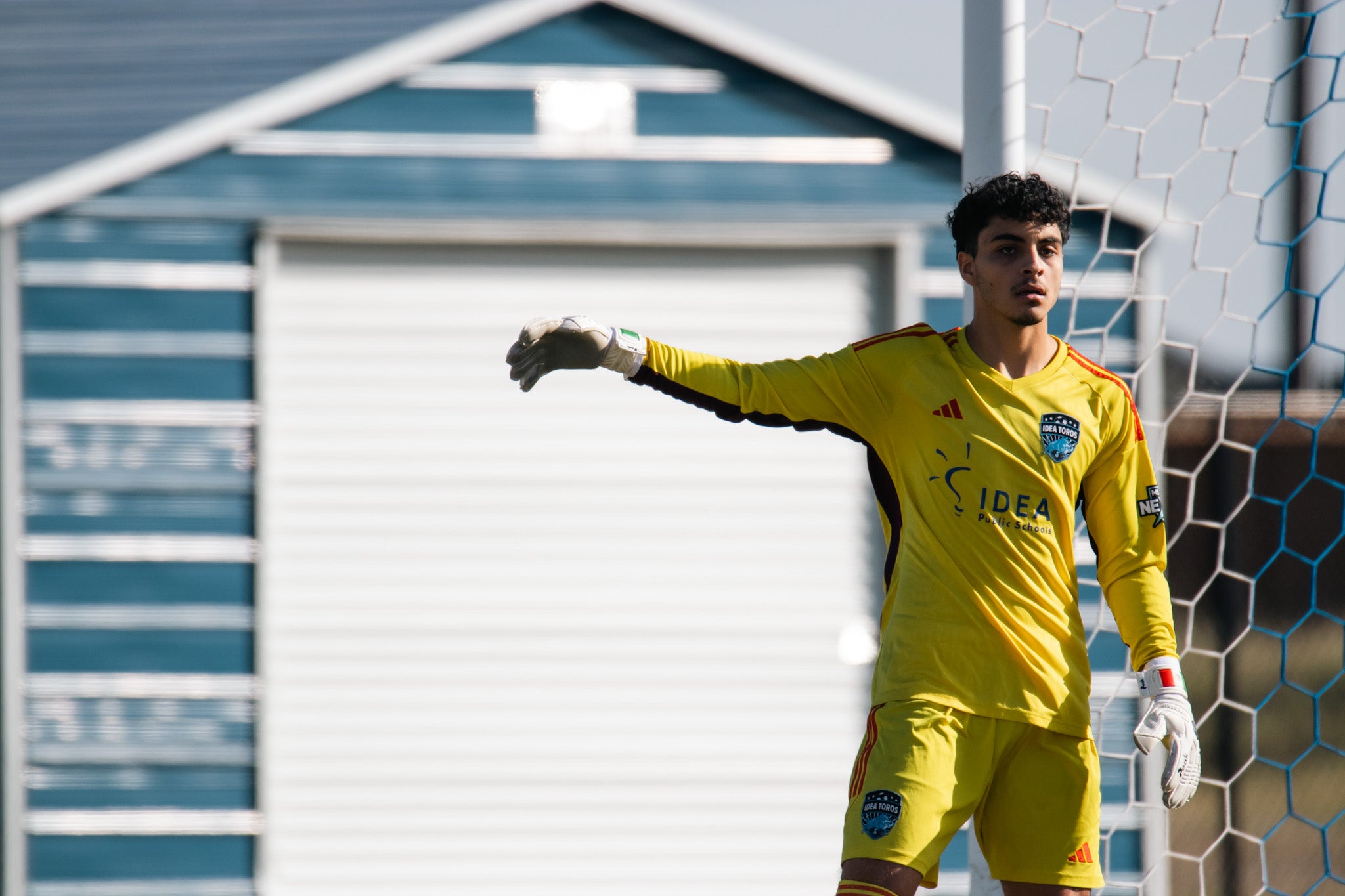 Soccer goalkeeper in yellow uniform standing in front of a goalpost with a beach hut in the background